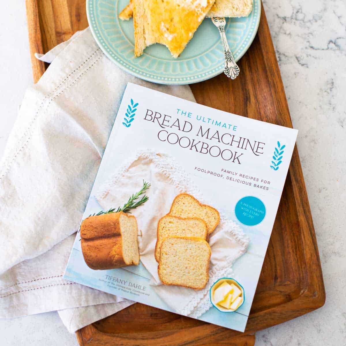 A copy of The Ultimate Bread Machine Cookbook sits on a wooden tray with a napkin and a plate of buttered toast.