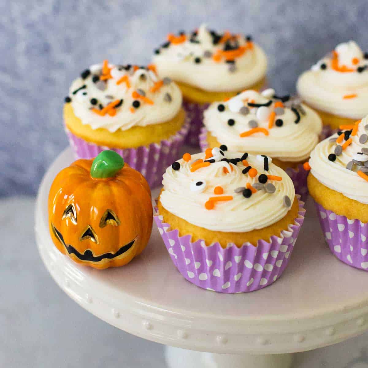 A cake plate has several vanilla cupcakes with orange and black sprinkles.