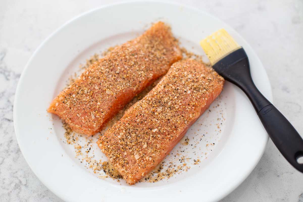The salmon fillets have been brushed with butter and sprinkled with seasoning. The basting brush sits on the plate next to them.