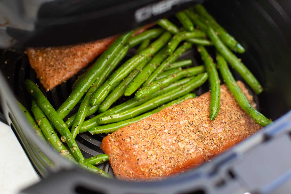 The salmon and green beans have been added to the air fryer basket in a single layer with the beans in the middle.