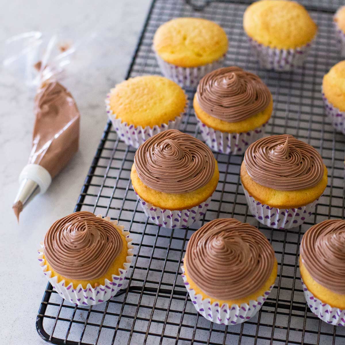 Cupcakes topped with a swirl of chocolate cream cheese frosting sit on a baker's rack next to a piping bag filled with frosting.