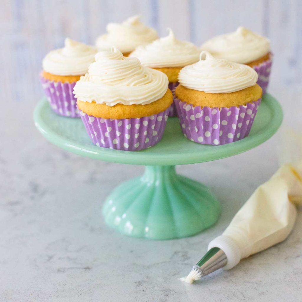 Vanilla cupcakes have been topped with a swirl of cream cheese frosting and sit on a cake plate. There's a frosting piping bag on the counter in front.