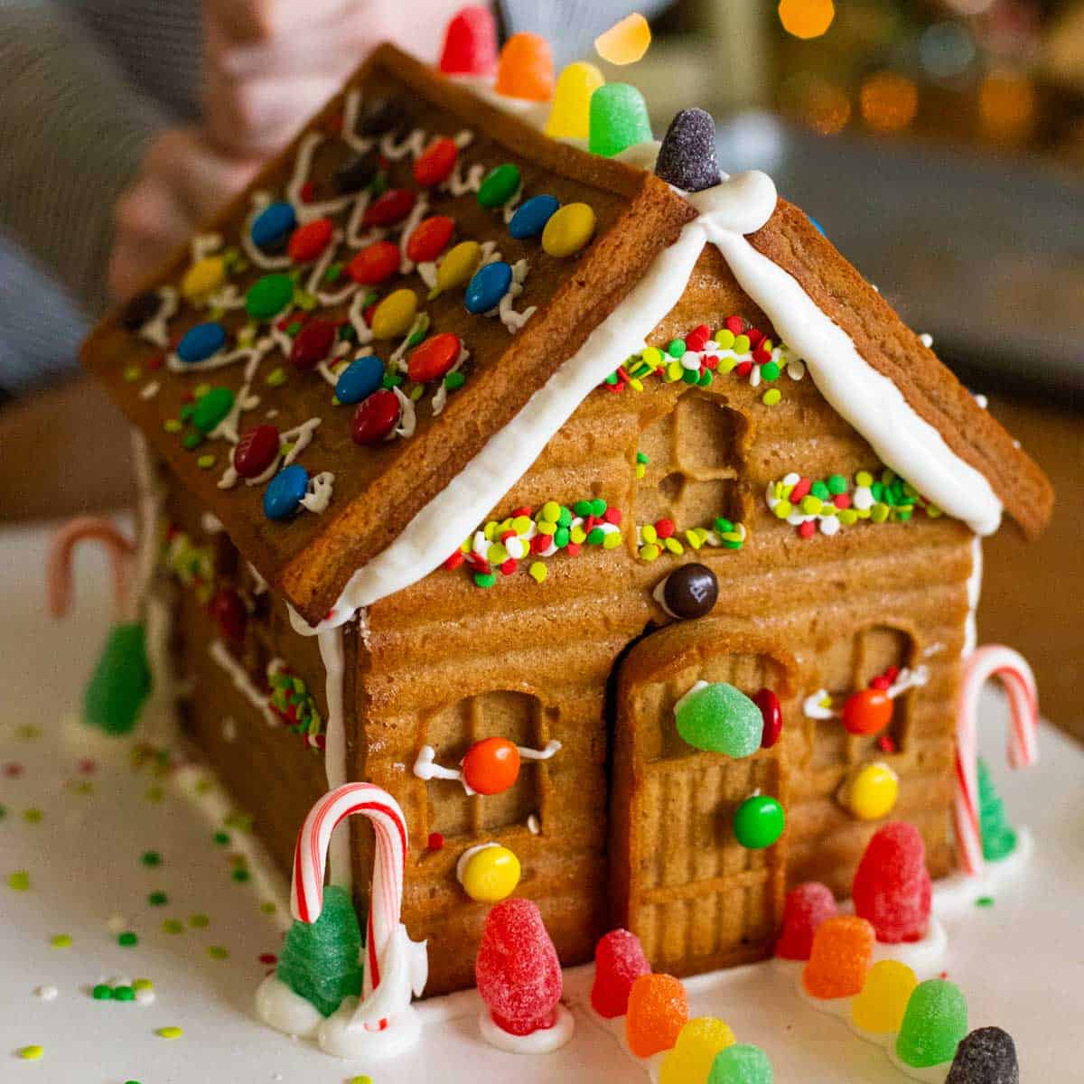 The finished gingerbread house has been assembled and decorated by a young girl with gumdrops, candy canes, and sprinkles.