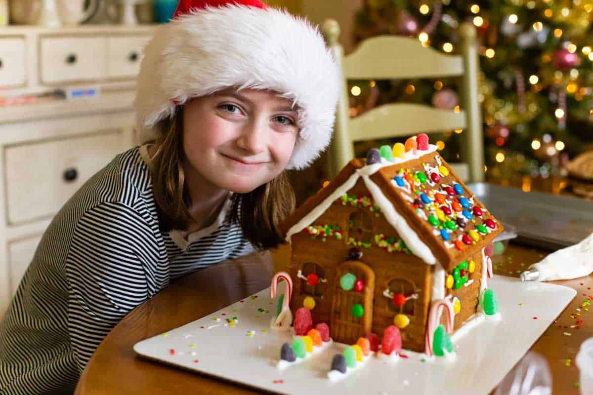 A young girl wearing a Santa hat shows off her homemade gingerbread house.