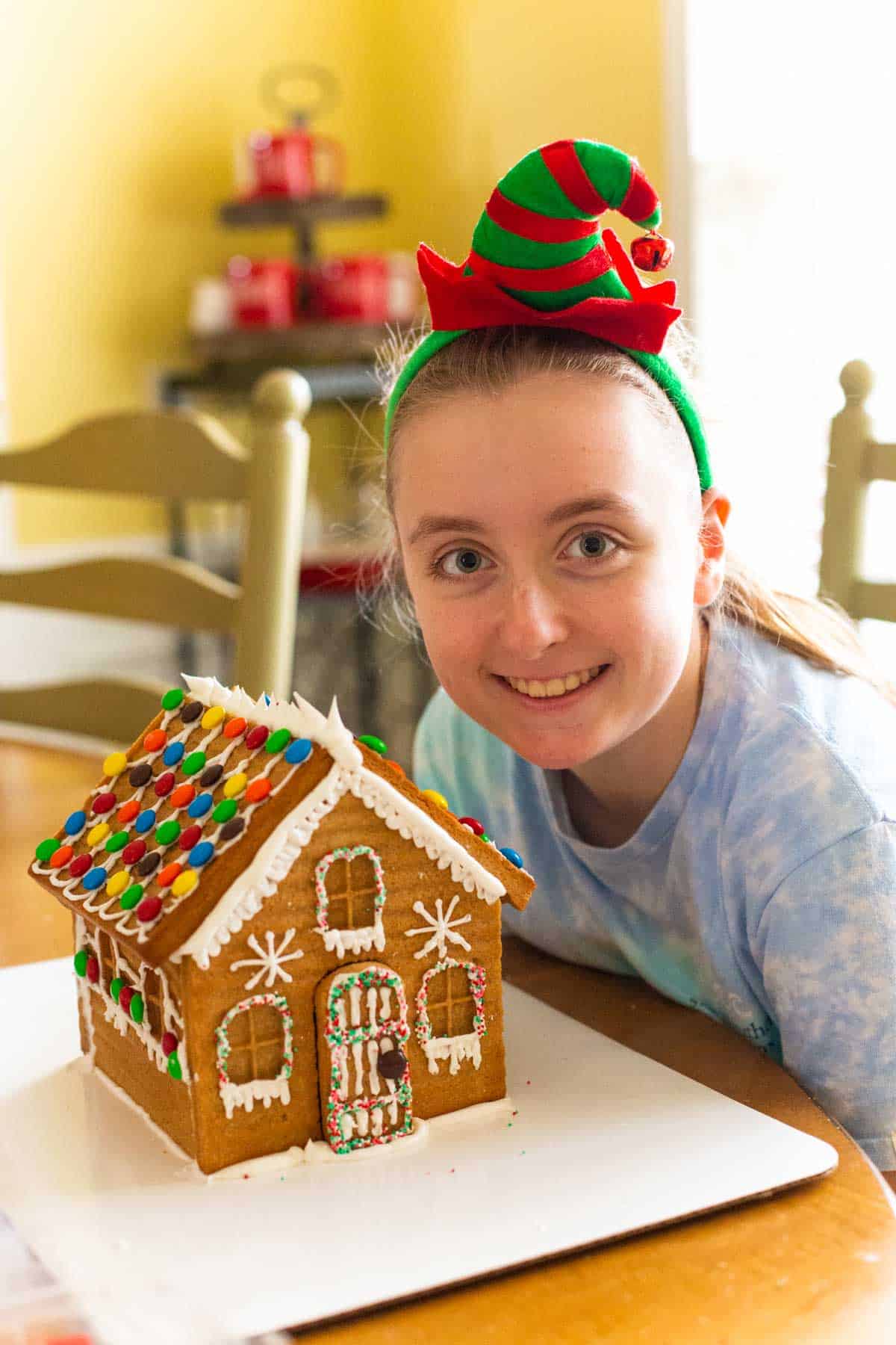 A teen girl wearing an elf headband shows off her finished gingerbread house.