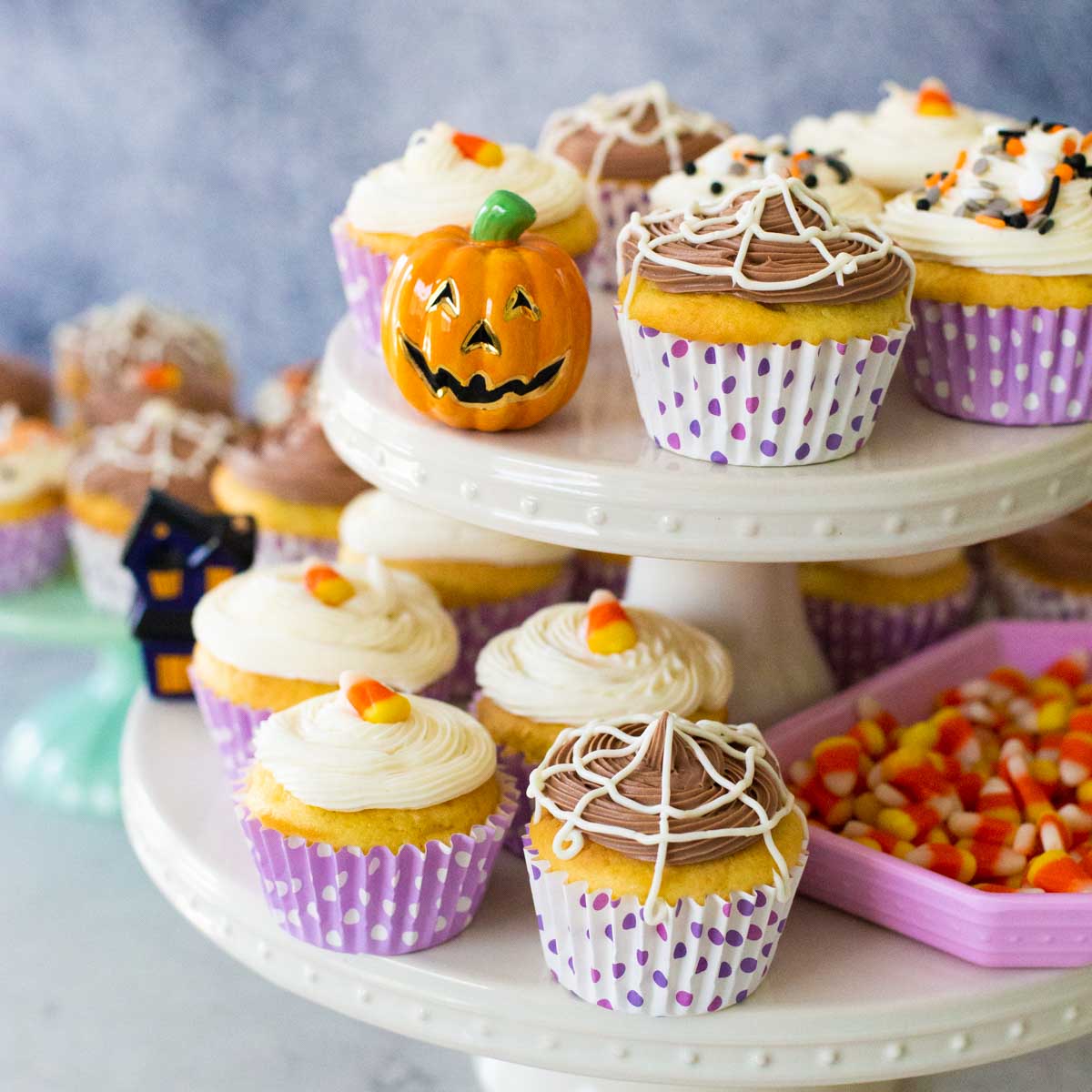 A two-tier cake plate has cute Halloween cupcakes in lavender polka dot wrappers. There is a dish of candy corn on the side.