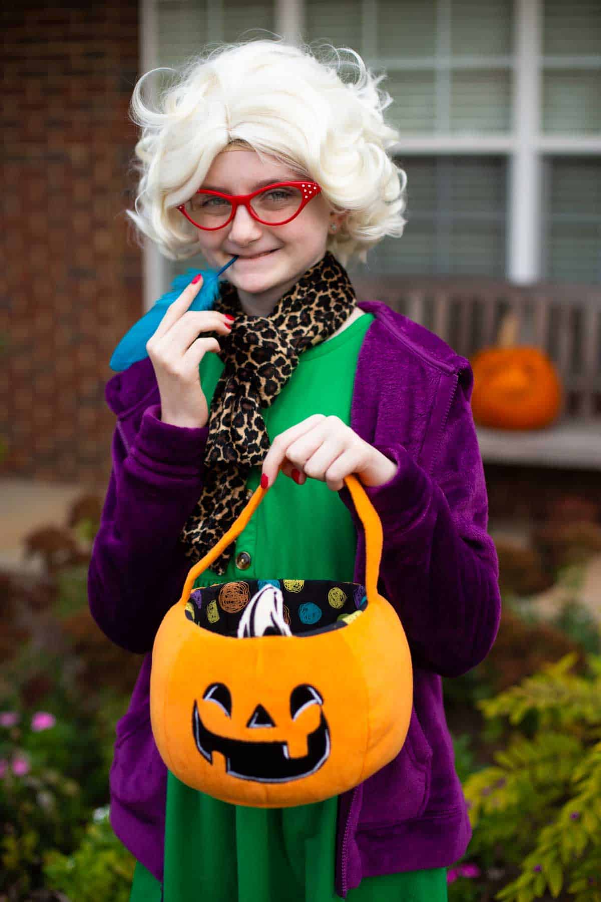 A young girl is wearing a Rita Skeeter wig, a green dress, a purple hoodie, and holding a pumpkin bag.