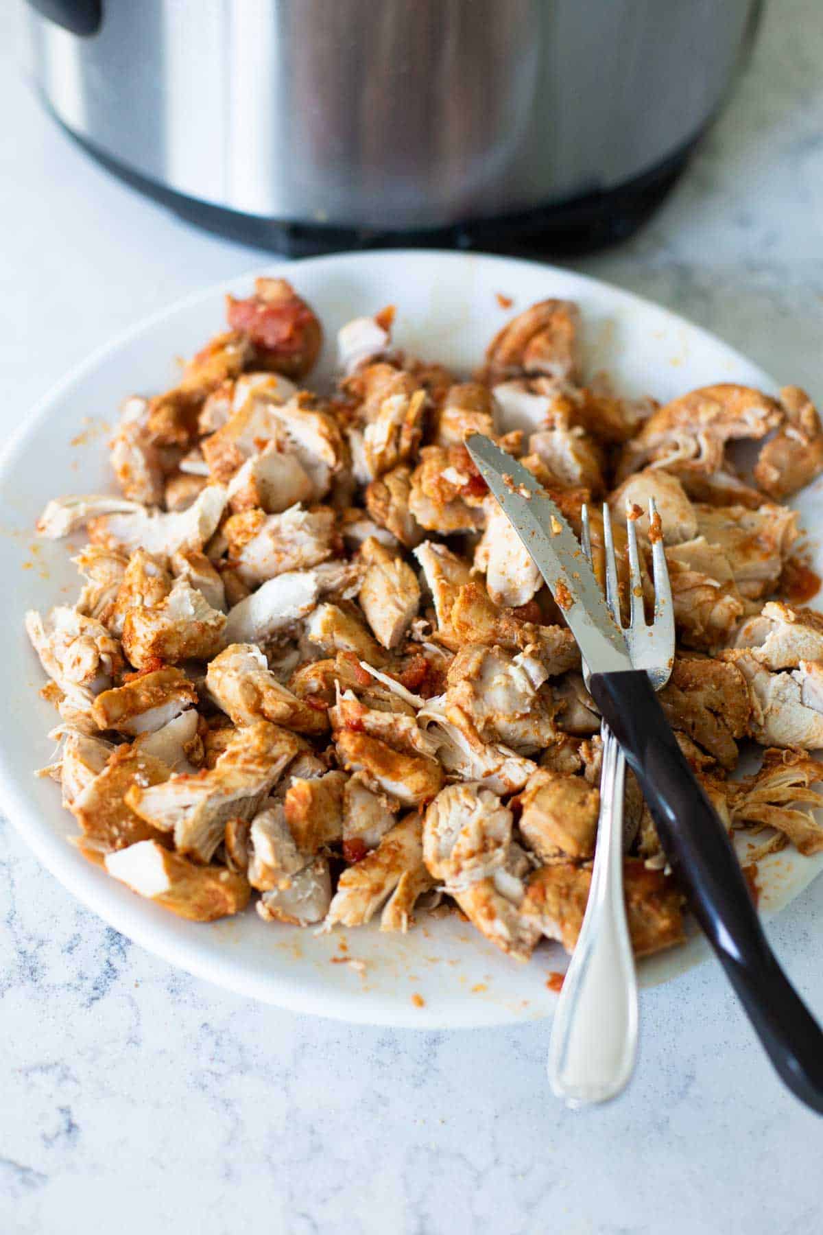 The chicken thighs have been cut with a knife and fork and are on a dinner plate next to the Instant Pot