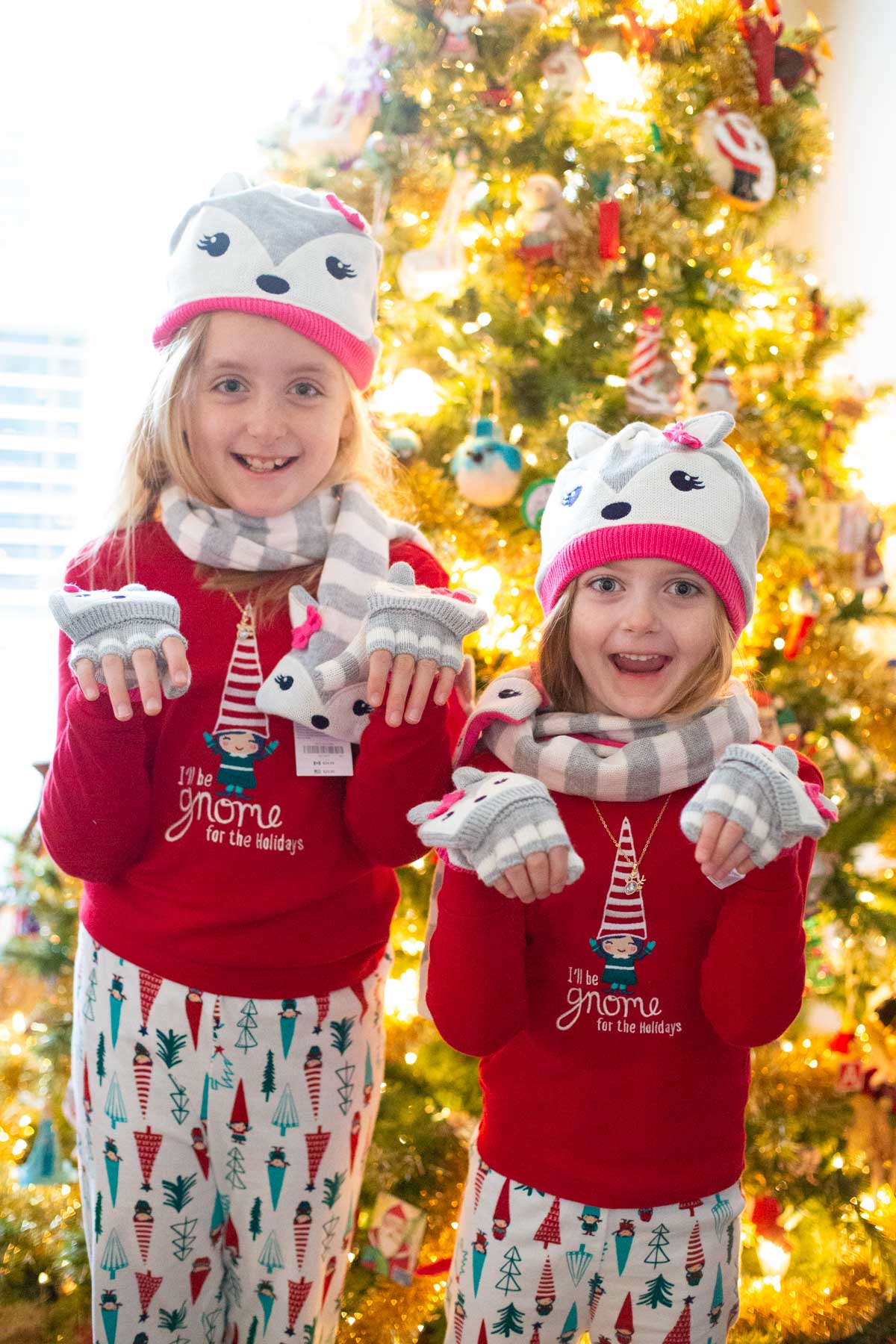 Two young girls wear brand new matching winter hats from St. Nick and are standing in front of a Christmas tree.