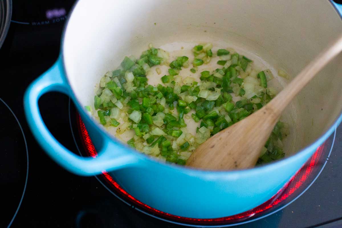 Chopped green pepper and onions are cooking in a soup pot with a wooden spoon.