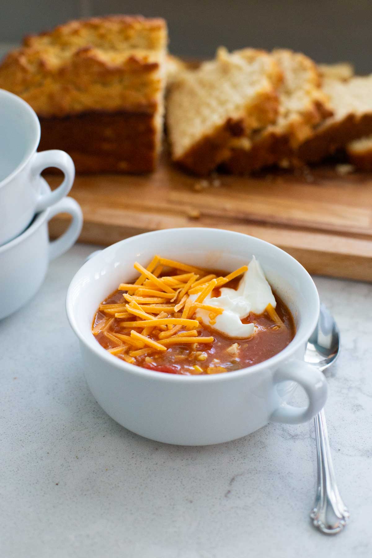 The taco soup is in a bowl with shredded cheese and a dollop of sour cream. Sliced bread is on a board in the back.