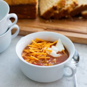 A bowl of hot chicken taco soup has shredded cheddar and a dollop of sour cream on top. Sliced bread in the background.