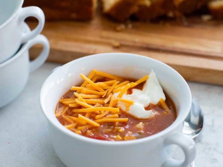 A bowl of hot chicken taco soup has shredded cheddar and a dollop of sour cream on top. Sliced bread in the background.