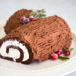 A chocolate and cream Christmas yule log cake roll with sugared cranberries sits on a cake platter.