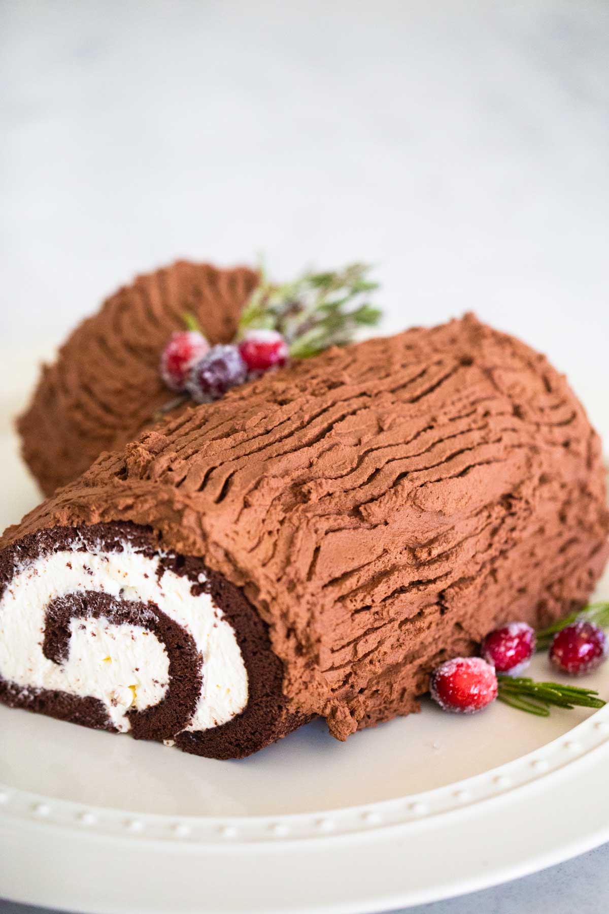 The finished yule log cake sits on a cake plate with sugared cranberries and rosemary.