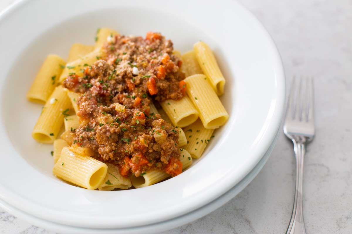 A white bowl filled with rigatoni pasta has been covered with a scoop of meaty bolognese sauce.
