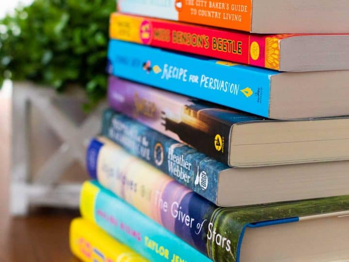 A stack of fiction books for book club in front of a green plant.
