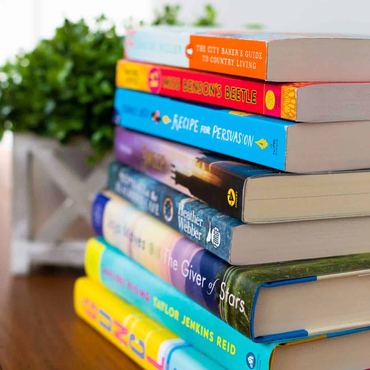 A stack of fiction books for book club in front of a green plant.