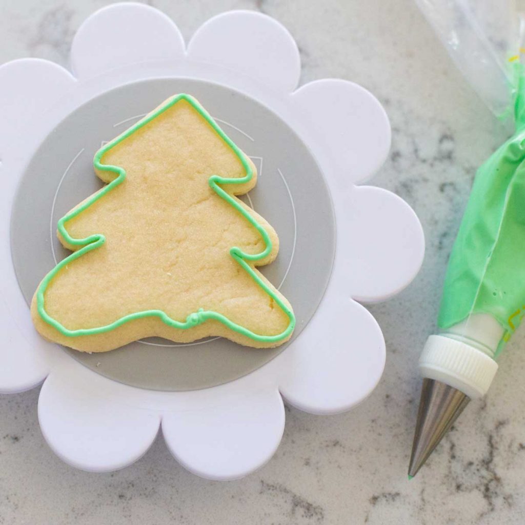A tree shaped sugar cookie has a green royal icing border that was piped on. The piping bag rests next to it.