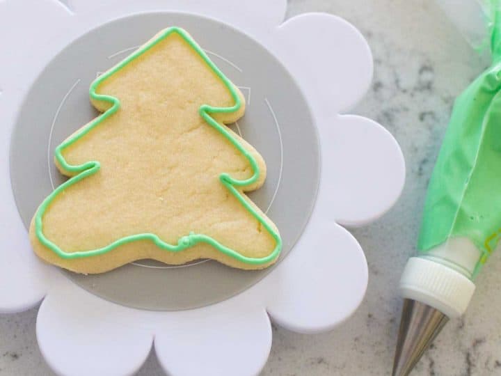 A tree shaped sugar cookie has a green royal icing border that was piped on. The piping bag rests next to it.