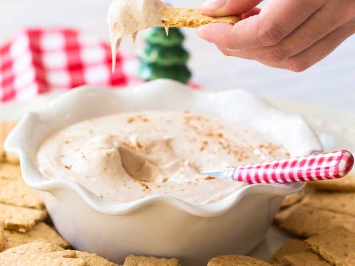 A hand is dunking a graham cracker into a creamy gingerbread dip.