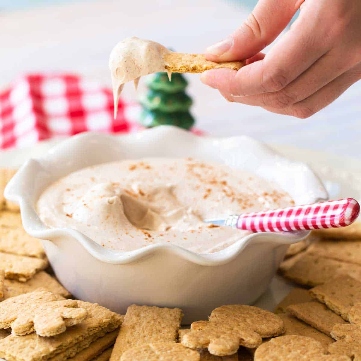 A hand is dunking a graham cracker into a creamy gingerbread dip. 