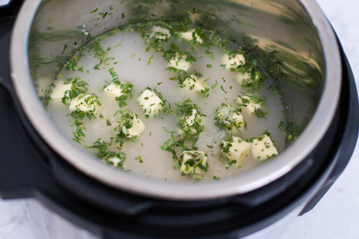 The herb butter rice ingredients have been stirred together in the instant pot. Butter cubes and herbs are floating on top of the water.