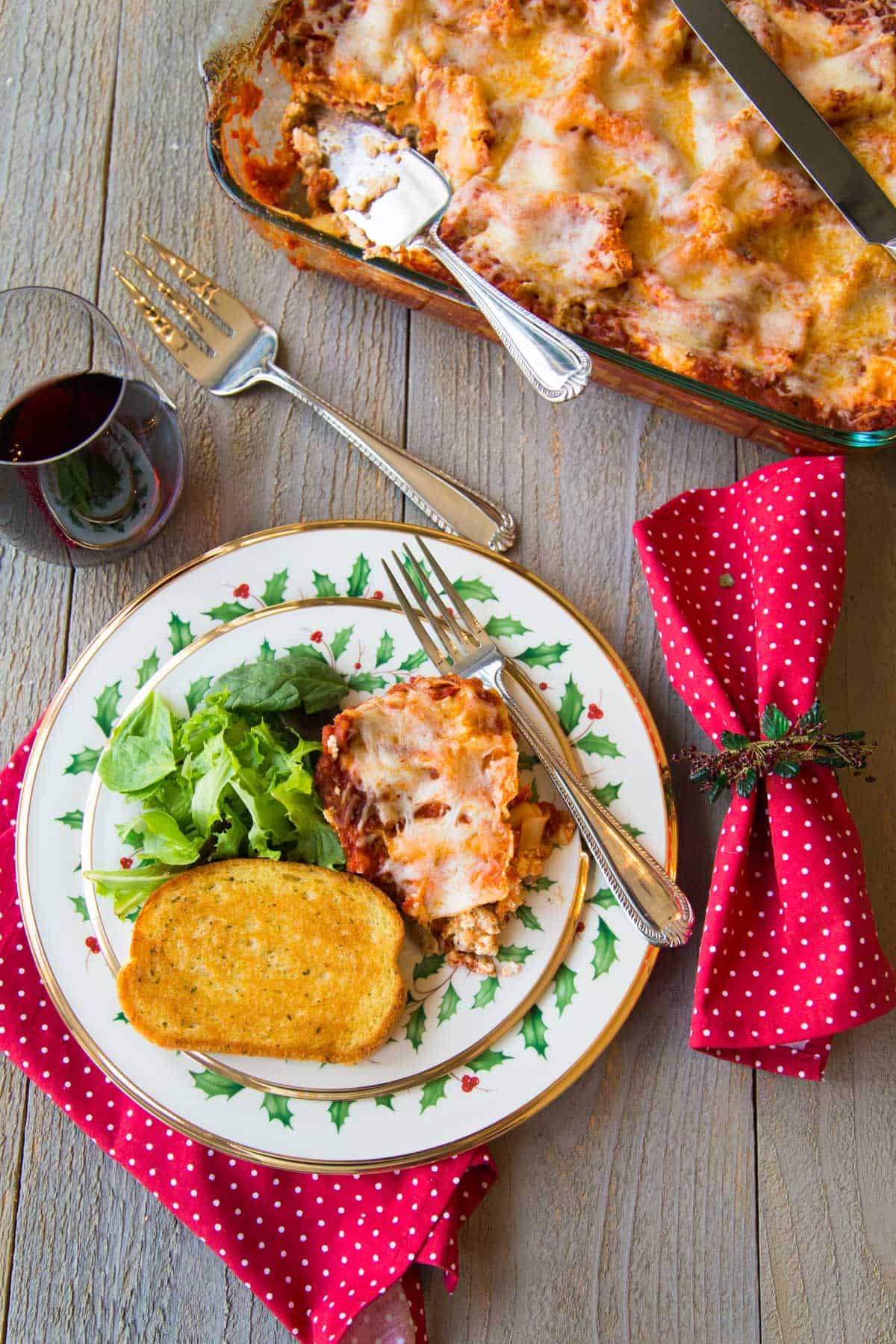 The finished casserole dish is on the table next to a festive Christmas china plate filled with a serving of lasagna, a piece of garlic bread, and a green salad.