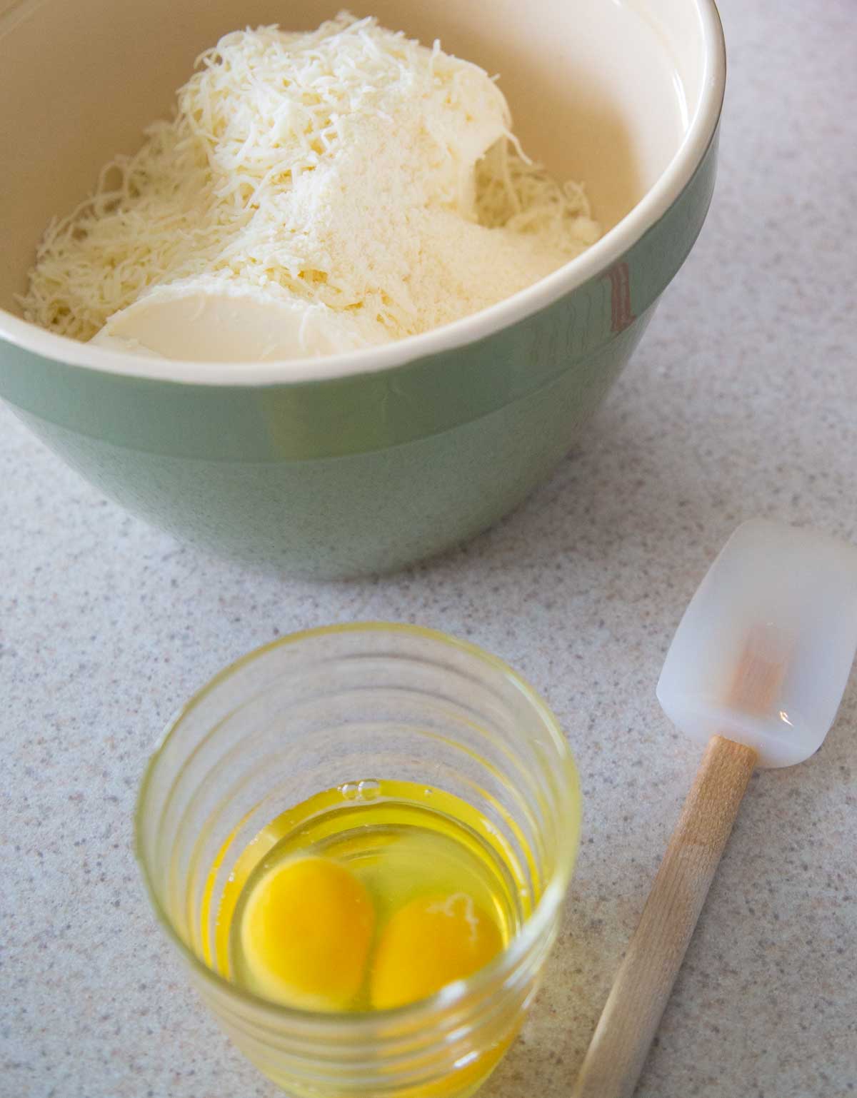 A bowl of eggs and another mixing bowl filled with ricotta and shredded cheese has a spatula ready to stir.