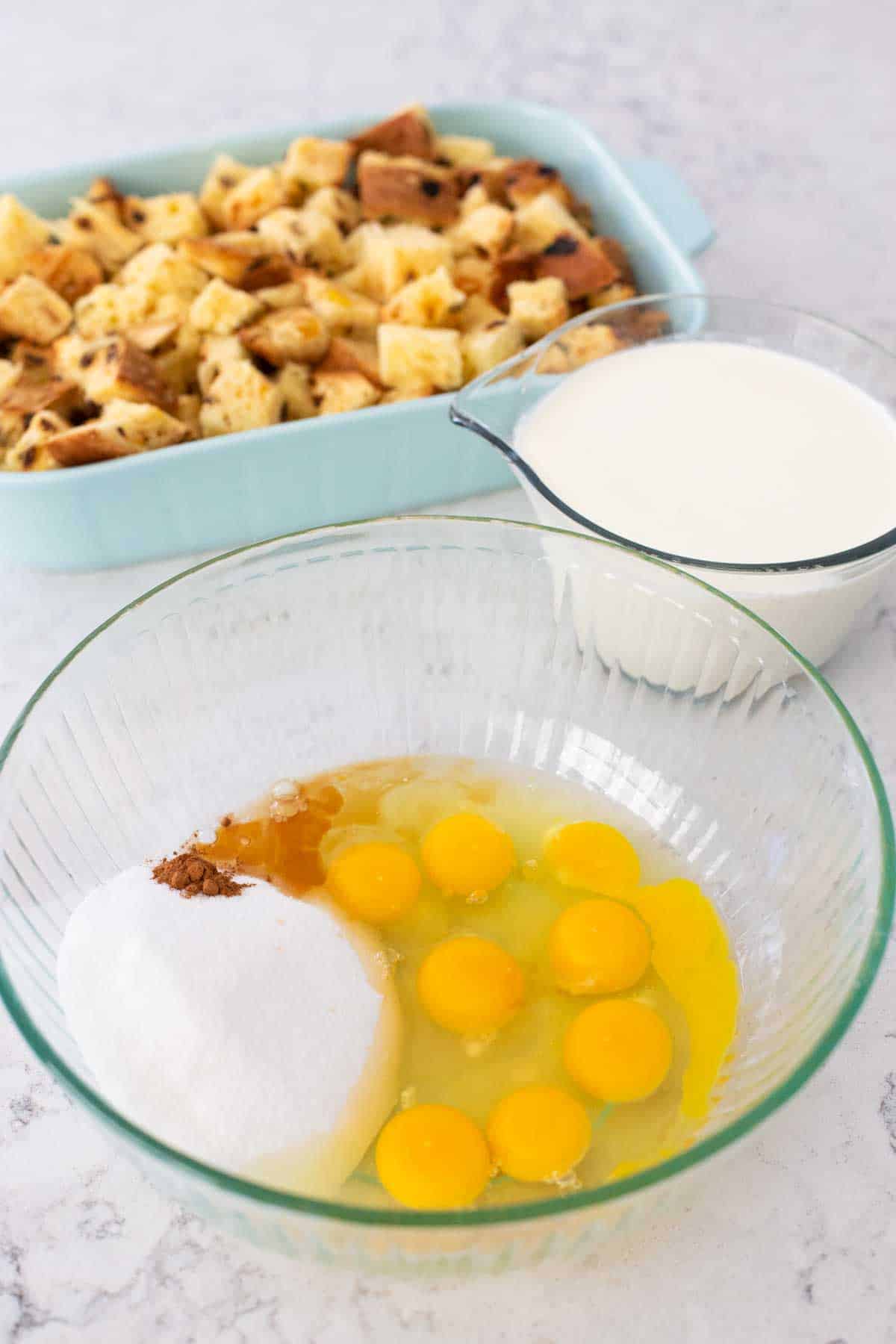 The eggs, sugar, and vanilla are in a large mixing bowl next to a measuring cup filled with milk and cream. The bread in the baking dish is in the background.