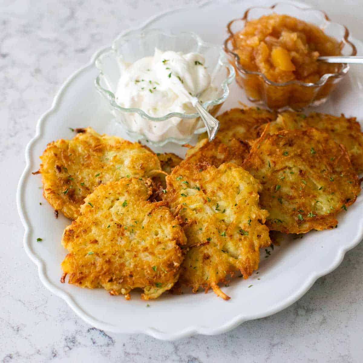 A platter of homemade potato latkes next to cups of sour cream and applesauce with spoons.