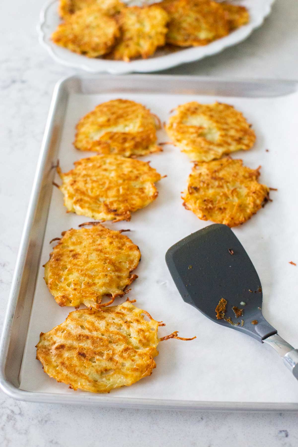 The latkes are lined up on a baking sheet with parchment paper and a spatula.