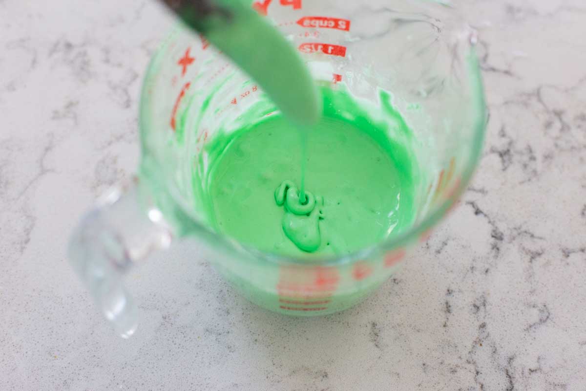 A measuring cup filled with green royal icing has a knife stirring it for consistency.