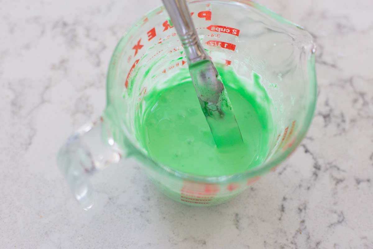 A measuring cup filled with green royal icing has a knife stirring it for consistency, it smooths over quickly.