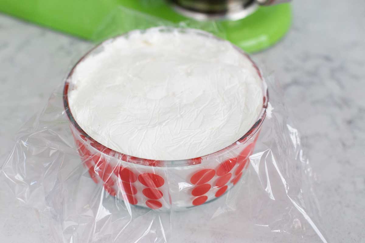 A glass storage container is filled to the brim with thick royal icing. Plastic wrap has been pressed over the whole surface before the lid goes on.