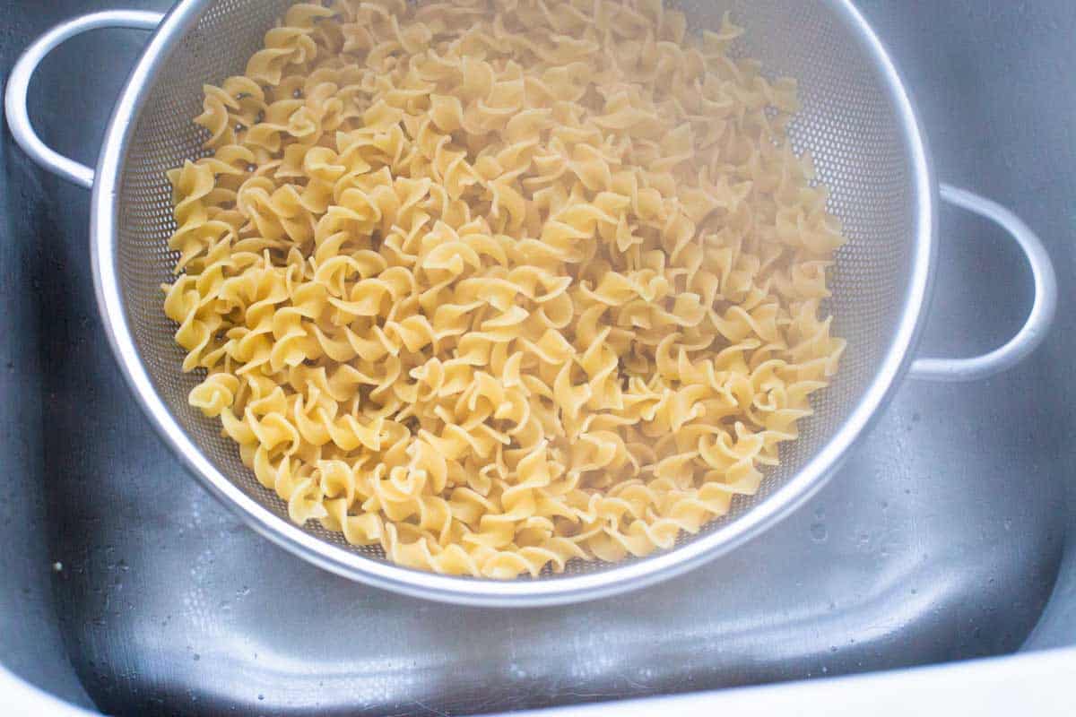 The wavy egg noodles have been boiled and drained in a strainer in the kitchen sink. Steam is rising up towards the camera.