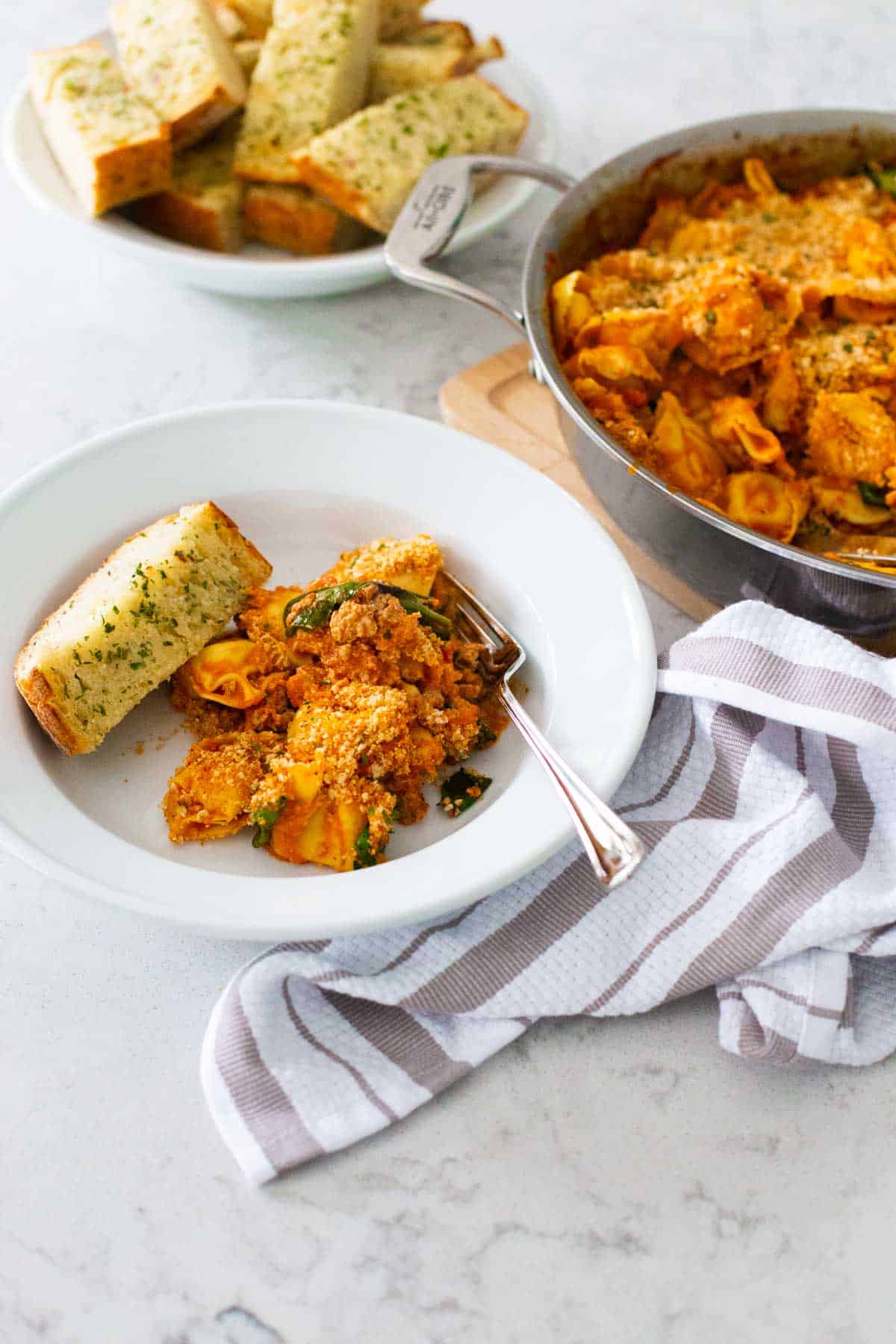 A serving of baked tortellini has a basket of garden bread nearby.