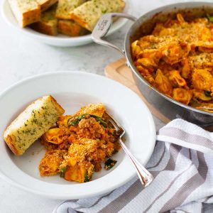 A bowl of baked tortellini with a slice of garlic bread on the side.