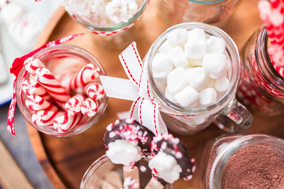 Hot chocolate station with variety of toppings including marshmallows, peppermint sticks, and chocolate coated spoons.