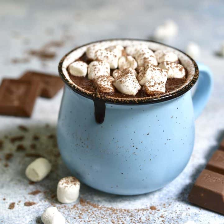 A blue mug filled with hot cocoa has mini marshmallows on top and chocolate pieces and marshmallows scattered around it.