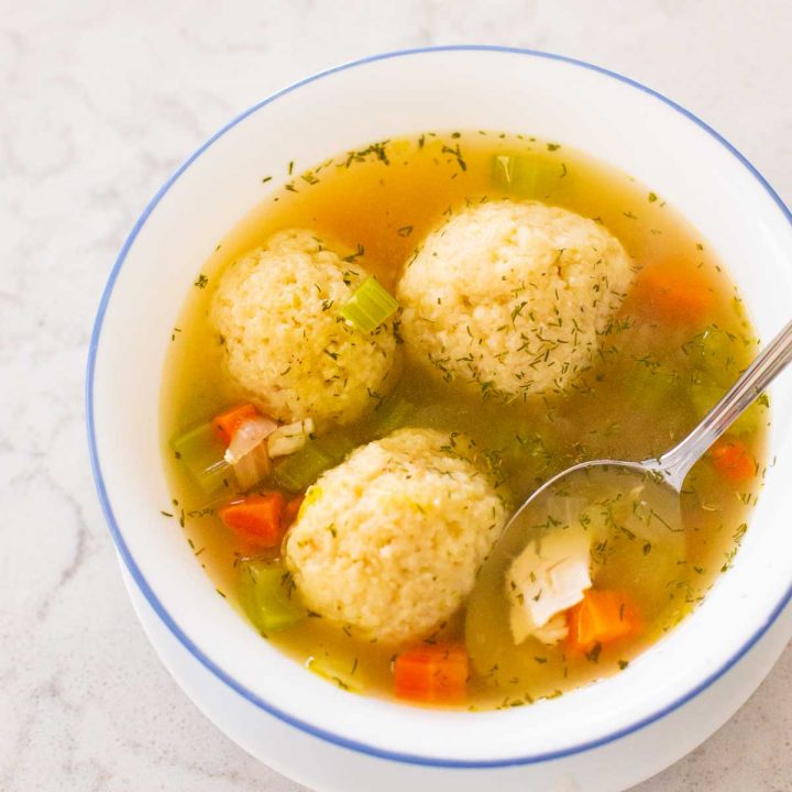 A bowl of homemade matzoh ball soup has a spoon showing the carrots and chicken while three matzoh balls float in the broth.