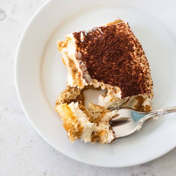 A slice of tiramisu is on a white plate, a spoon has a bite on it showing the cream filling and spongey cookie layer.