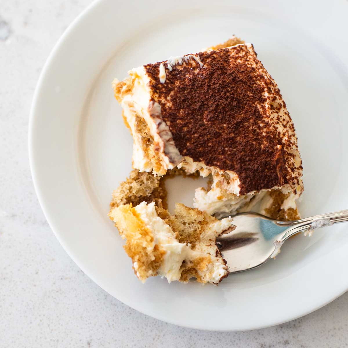 A slice of tiramisu is on a white plate, a spoon has a bite on it showing the cream filling and spongey cookie layer.