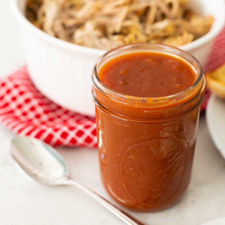A mason jar filled with barbecue sauce has a spoon and is in front of a dish of shredded pork.