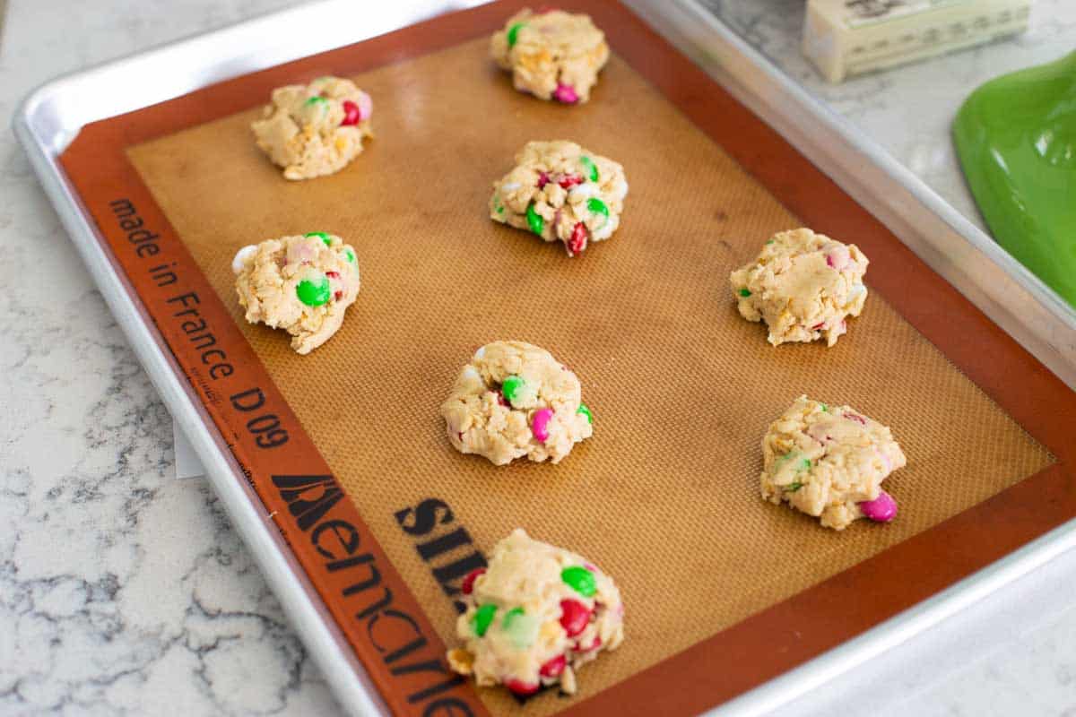 Portions of cookie dough have been placed on a baking sheet lined with a mat.