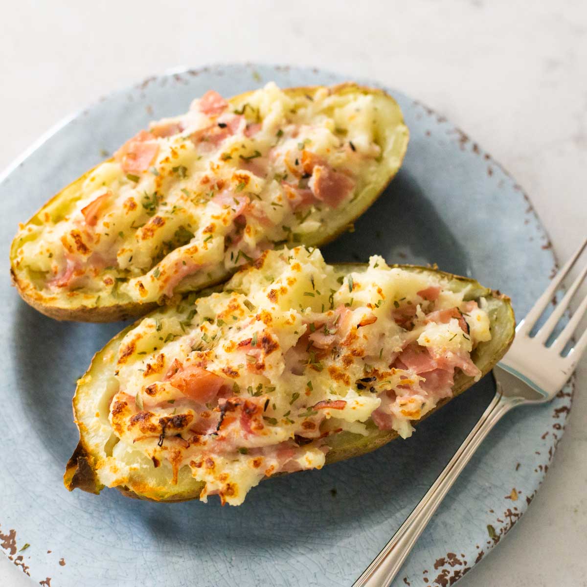 Two ham and cheese stuffed potatoes are on a serving plate with a fork.