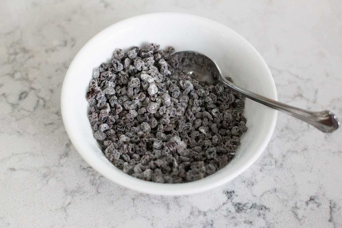 A bowl of dried currants have been stirred together with flour with a spoon.