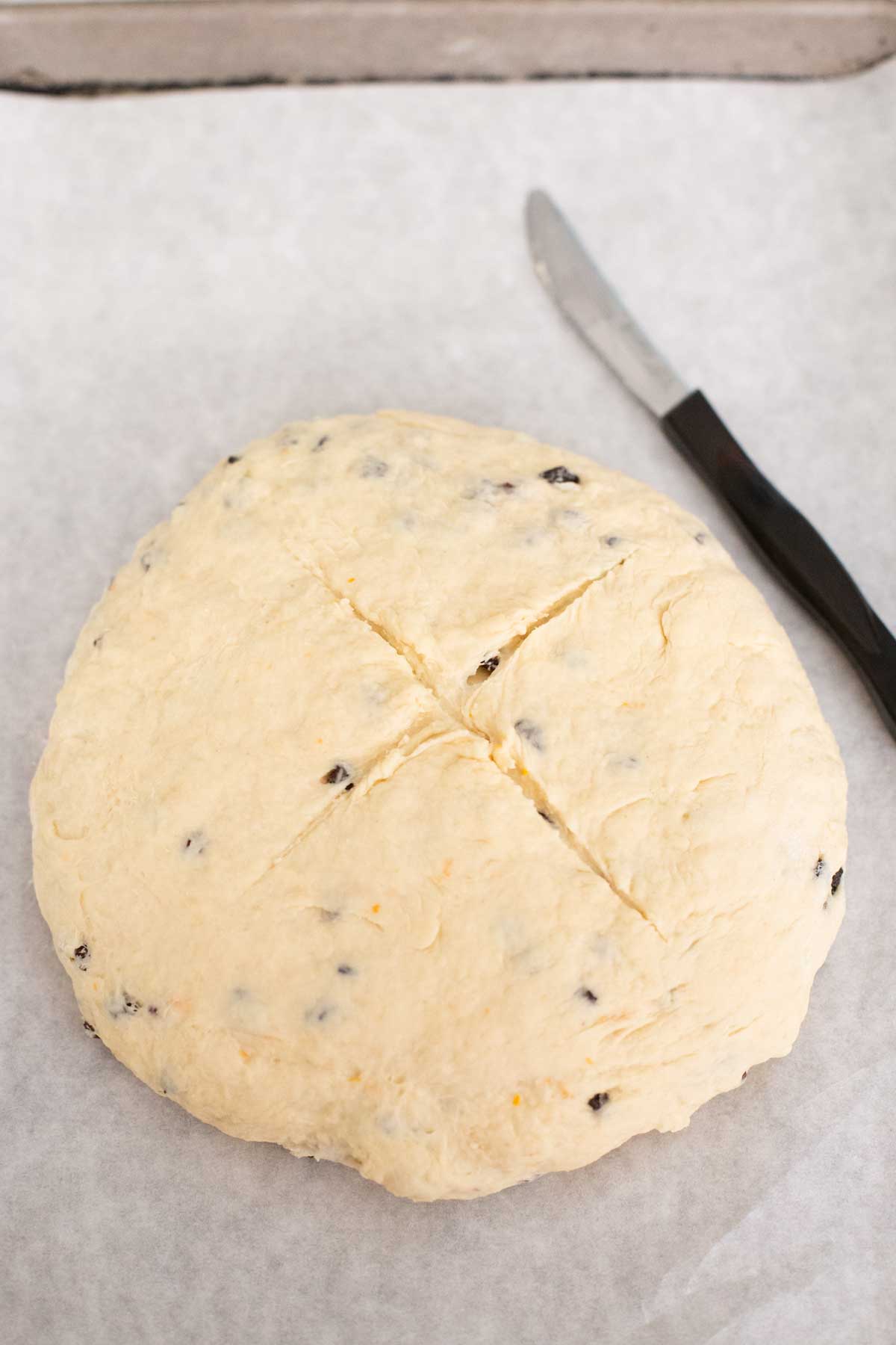 The dough has been shaped into a round disk and a cross has been sliced on the top with a small knife that sits on the tray next to it.