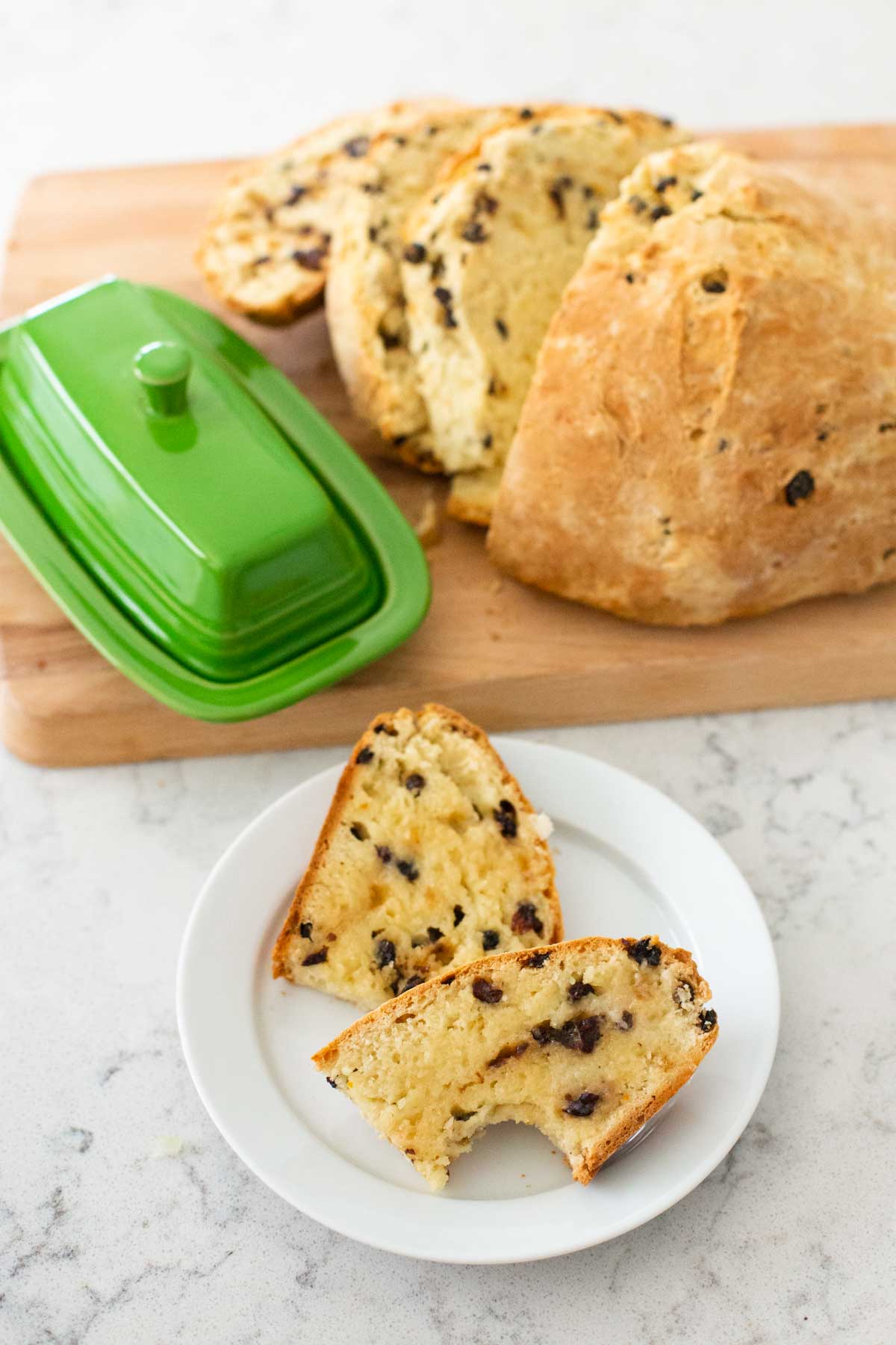 The Irish soda bread has been sliced and served with a pat of butter on a small plate. The butter dish sits on the cutting board next to the loaf.