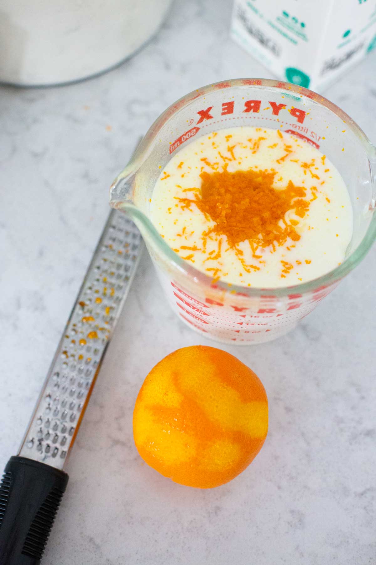 A measuring cup of buttermilk has fresh orange zest grated over the top and the zesting tool to the side.